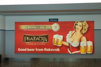 Vyprsená Pražačka s pivem v ruce vítá turisty: Kontroverzní reklama na letišti v Praze dál zůstává